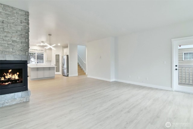 unfurnished living room with a stone fireplace, stairway, light wood-style flooring, and baseboards