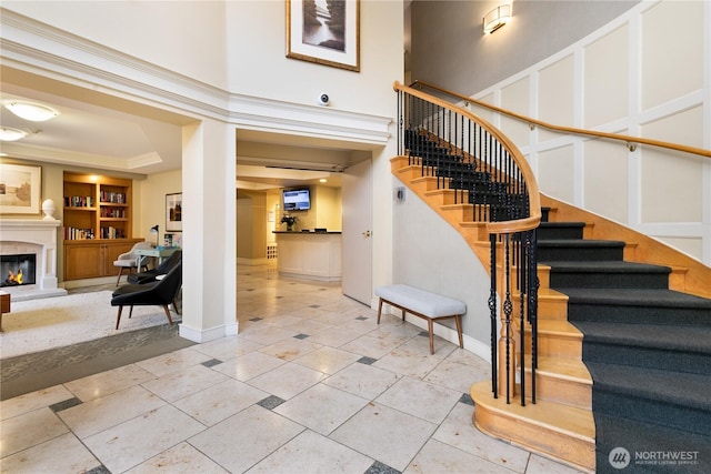 staircase with built in features, a towering ceiling, ornamental molding, a glass covered fireplace, and tile patterned floors