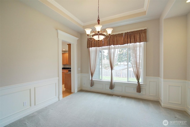 unfurnished dining area featuring visible vents, light colored carpet, a tray ceiling, wainscoting, and a notable chandelier