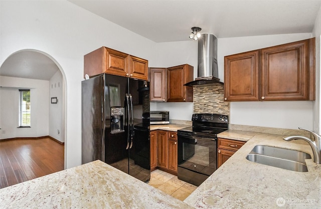 kitchen with light stone counters, a sink, wall chimney range hood, brown cabinets, and black appliances