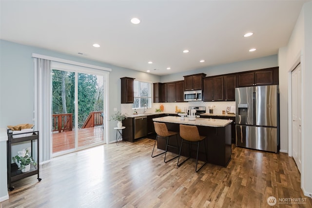 kitchen with wood finished floors, a kitchen island, dark brown cabinets, appliances with stainless steel finishes, and decorative backsplash