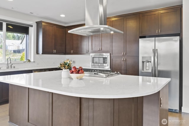 kitchen with island exhaust hood, appliances with stainless steel finishes, a sink, a kitchen island, and light stone countertops