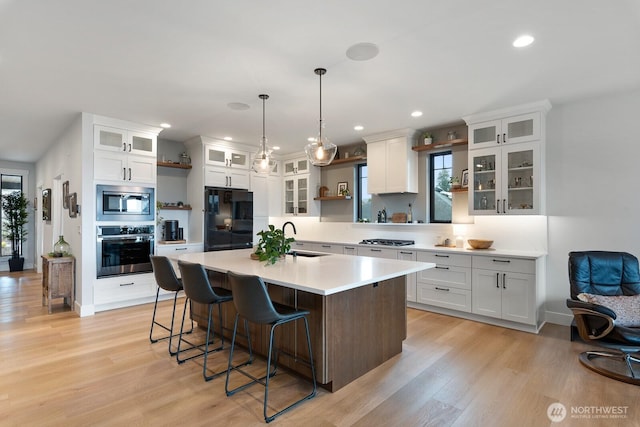 kitchen with light wood finished floors, open shelves, light countertops, appliances with stainless steel finishes, and a kitchen island with sink