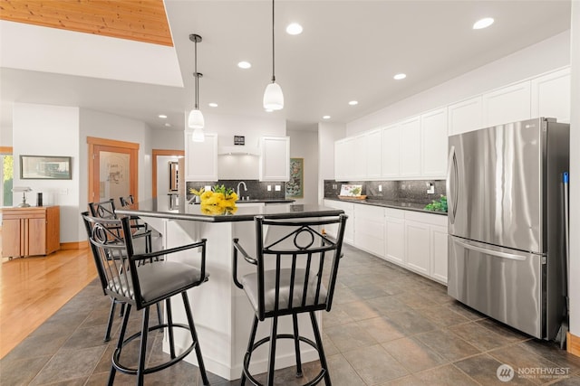 kitchen featuring a breakfast bar, backsplash, freestanding refrigerator, dark countertops, and a center island with sink