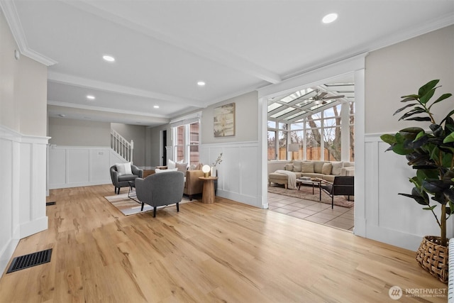 living room featuring light wood-type flooring, visible vents, beamed ceiling, and wainscoting