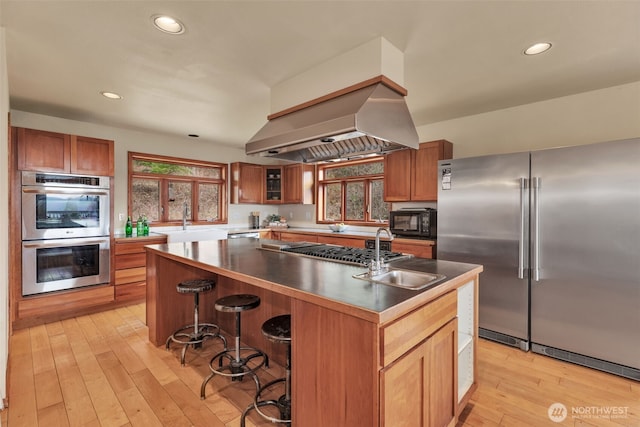 kitchen featuring island exhaust hood, appliances with stainless steel finishes, brown cabinetry, a sink, and light wood-type flooring