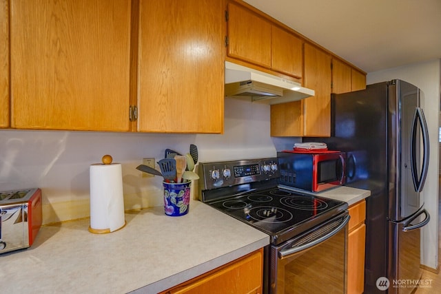 kitchen featuring under cabinet range hood, light countertops, freestanding refrigerator, stainless steel electric range oven, and brown cabinetry