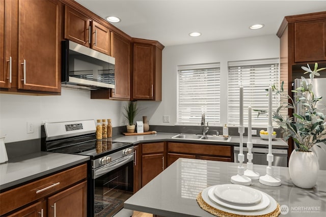 kitchen with brown cabinetry, dark countertops, stainless steel appliances, a sink, and recessed lighting