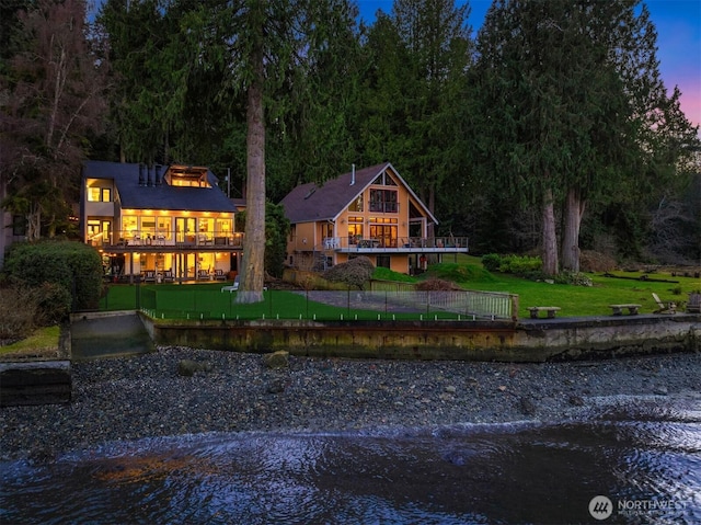 back of property at dusk featuring a water view, a lawn, a balcony, and fence