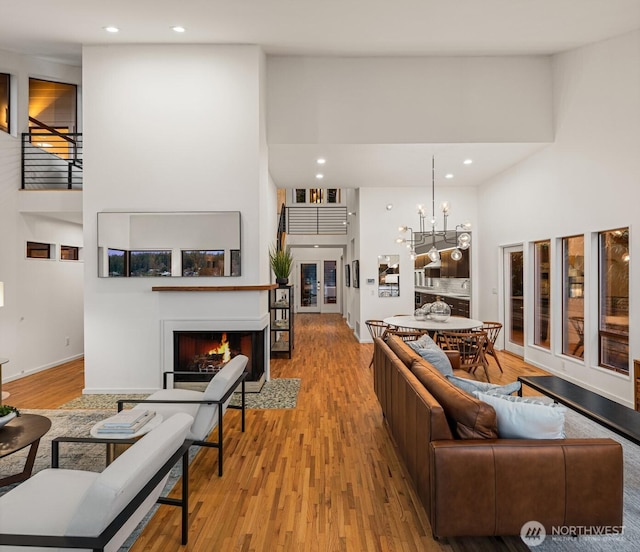 living area featuring light wood-style flooring, recessed lighting, a notable chandelier, a fireplace with flush hearth, and a towering ceiling