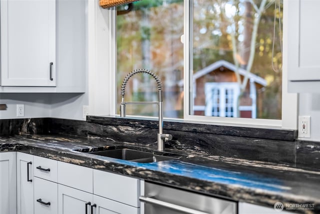 kitchen featuring stainless steel dishwasher, dark stone countertops, a sink, and white cabinets