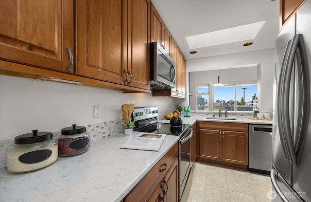 kitchen featuring a skylight, stainless steel appliances, brown cabinetry, a sink, and light stone countertops