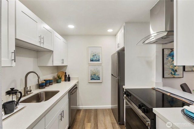 kitchen with light countertops, appliances with stainless steel finishes, white cabinets, a sink, and wall chimney range hood