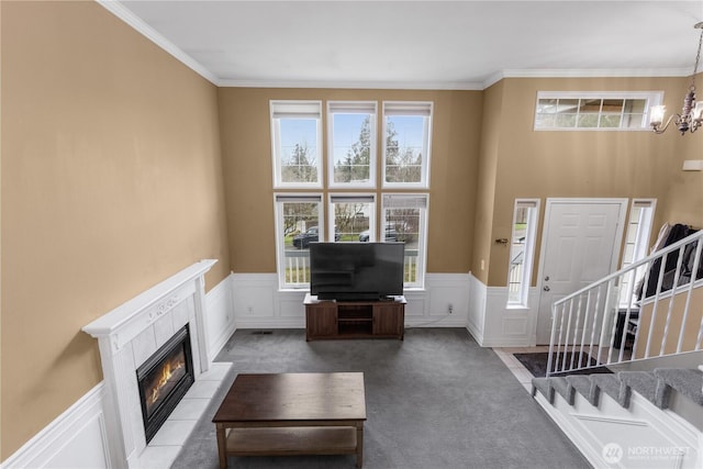 carpeted living area featuring a tiled fireplace, stairway, a notable chandelier, and wainscoting