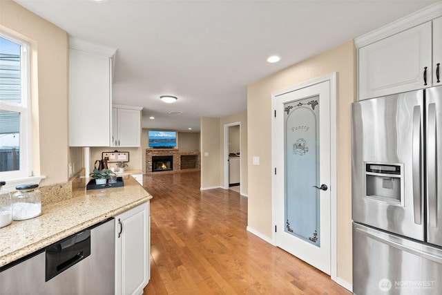 kitchen featuring light wood finished floors, appliances with stainless steel finishes, light stone countertops, a brick fireplace, and white cabinetry