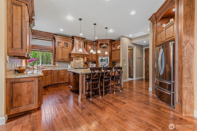 kitchen featuring a breakfast bar area, custom exhaust hood, appliances with stainless steel finishes, brown cabinetry, and a kitchen island