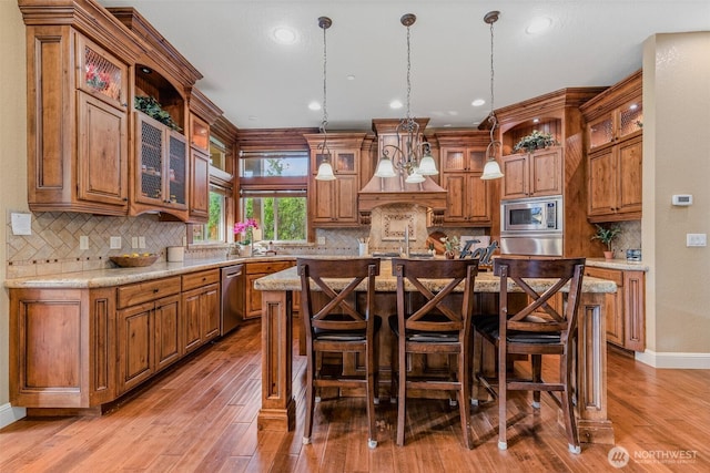 kitchen with stainless steel appliances, an island with sink, brown cabinetry, glass insert cabinets, and pendant lighting