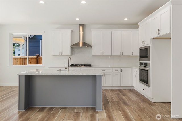 kitchen with oven, built in microwave, decorative backsplash, wall chimney exhaust hood, and an island with sink