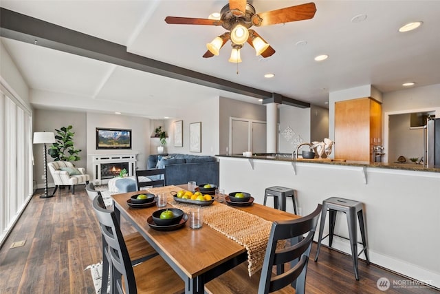 dining room featuring baseboards, recessed lighting, a lit fireplace, dark wood-type flooring, and beamed ceiling