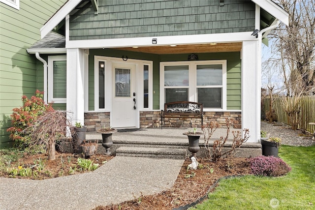 property entrance featuring covered porch, stone siding, a shingled roof, and fence