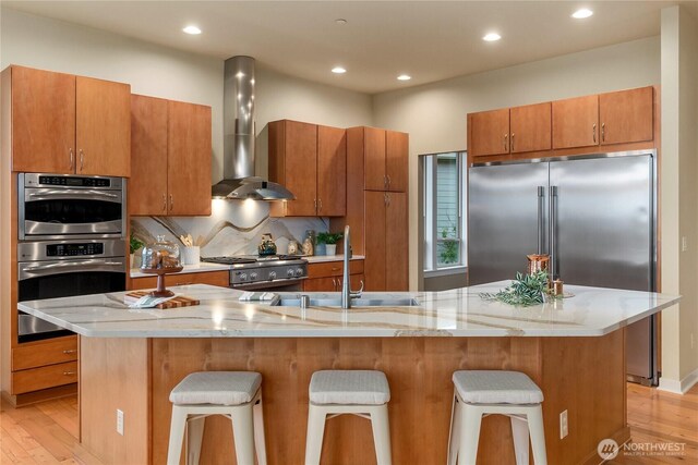 kitchen with wall chimney range hood, a kitchen breakfast bar, backsplash, brown cabinetry, and light wood finished floors