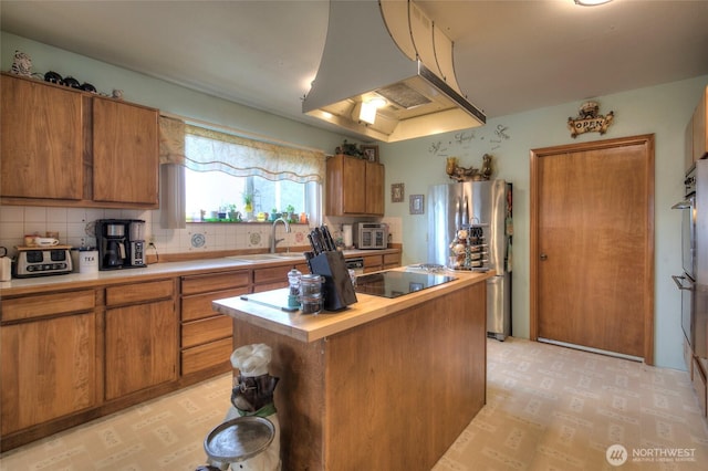 kitchen with black electric cooktop, exhaust hood, a sink, freestanding refrigerator, and brown cabinetry