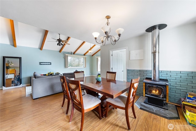 dining room featuring lofted ceiling with beams, ceiling fan with notable chandelier, light wood-type flooring, and a wood stove