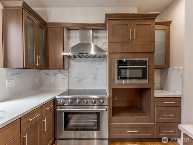 kitchen featuring tasteful backsplash, wall chimney exhaust hood, glass insert cabinets, appliances with stainless steel finishes, and light stone counters