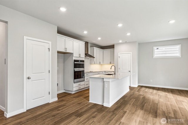 kitchen with dark wood-style flooring, a sink, a kitchen island with sink, double oven, and backsplash