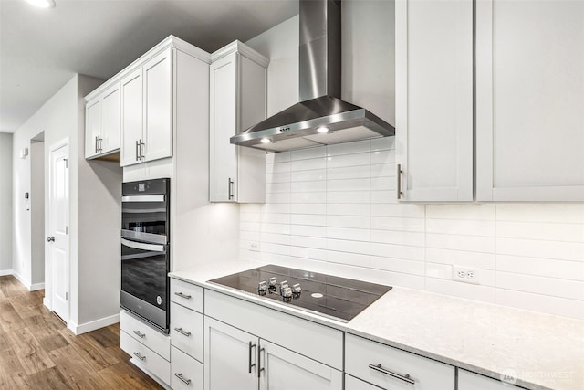 kitchen with wood finished floors, multiple ovens, black electric stovetop, wall chimney range hood, and backsplash