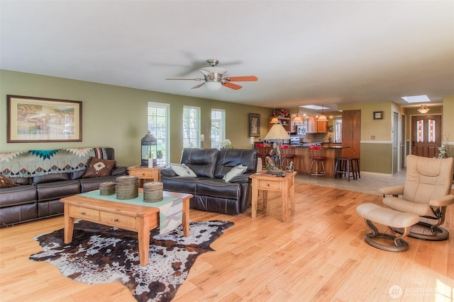 living area with light wood-style floors, ceiling fan, a skylight, and a wainscoted wall