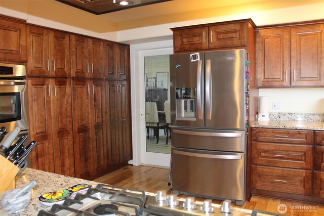 kitchen with appliances with stainless steel finishes, brown cabinetry, light wood-type flooring, and light stone counters