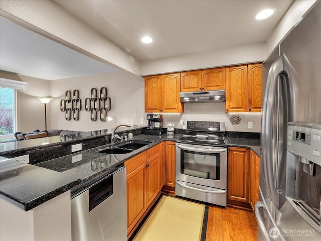 kitchen with dark stone counters, a peninsula, stainless steel appliances, under cabinet range hood, and a sink