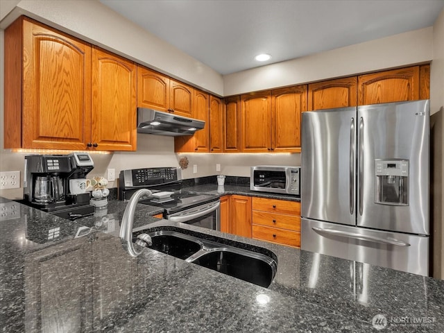 kitchen featuring under cabinet range hood, stainless steel appliances, a sink, brown cabinetry, and dark stone countertops