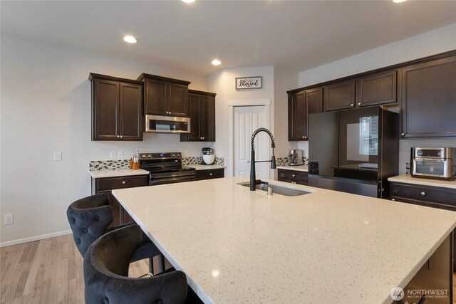 kitchen with a center island with sink, stainless steel appliances, a sink, dark brown cabinets, and a kitchen breakfast bar