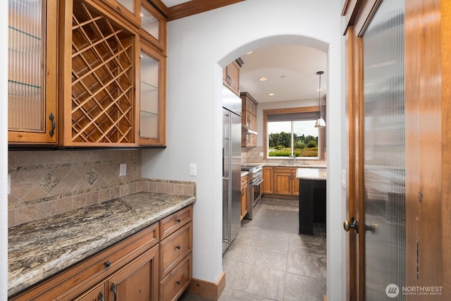 kitchen with light stone counters, arched walkways, high end appliances, backsplash, and brown cabinetry