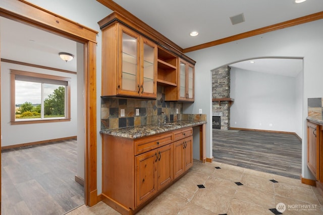 kitchen featuring arched walkways, decorative backsplash, ornamental molding, glass insert cabinets, and dark stone counters