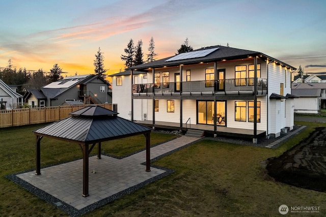 back of house at dusk with a balcony, a yard, a gazebo, a patio area, and roof mounted solar panels