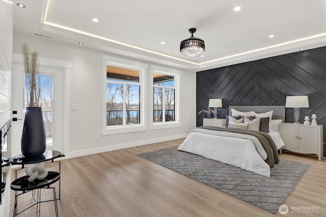 bedroom with an accent wall, light wood-type flooring, a raised ceiling, and baseboards