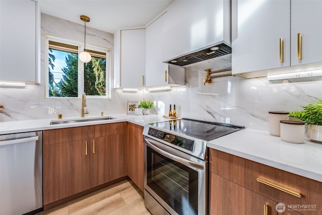 kitchen featuring pendant lighting, appliances with stainless steel finishes, brown cabinetry, a sink, and wall chimney exhaust hood