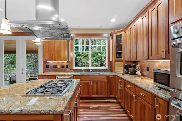 kitchen featuring brown cabinets, island exhaust hood, appliances with stainless steel finishes, a sink, and light stone countertops