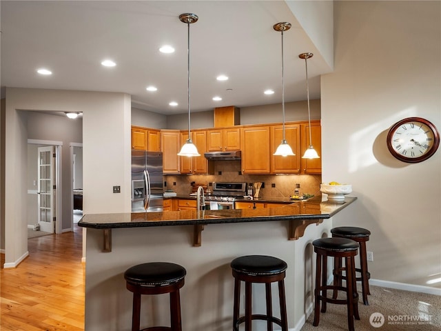 kitchen with decorative backsplash, appliances with stainless steel finishes, brown cabinetry, under cabinet range hood, and a kitchen bar