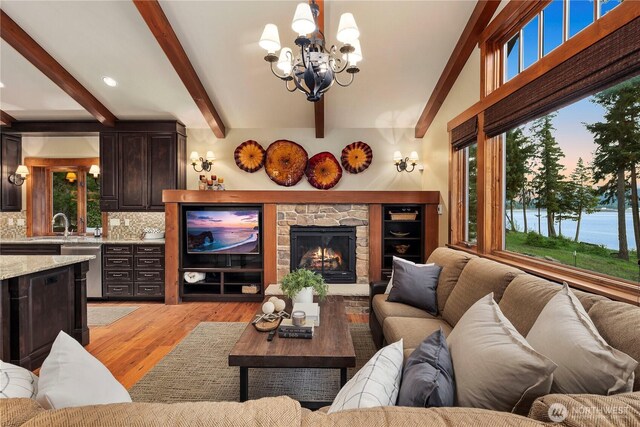 living area featuring light wood finished floors, beam ceiling, a notable chandelier, and a stone fireplace