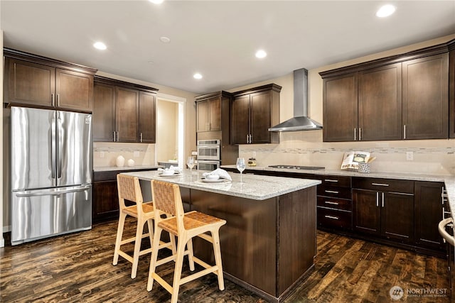 kitchen featuring light stone counters, dark brown cabinetry, stainless steel appliances, wall chimney range hood, and dark wood finished floors