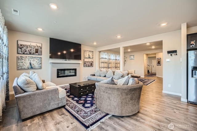 living room featuring recessed lighting, visible vents, light wood-style floors, a glass covered fireplace, and baseboards