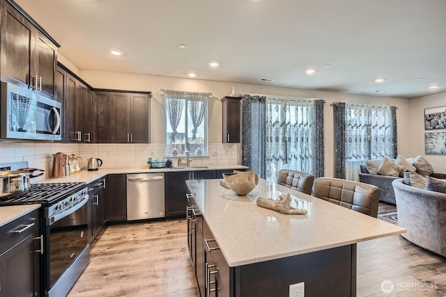 kitchen with stainless steel appliances, light wood-style flooring, open floor plan, dark brown cabinets, and light stone countertops