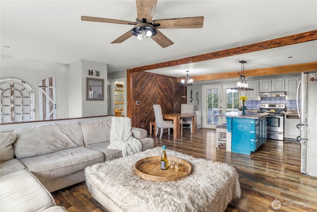 living room with dark wood-style floors, beamed ceiling, wood walls, and ceiling fan with notable chandelier