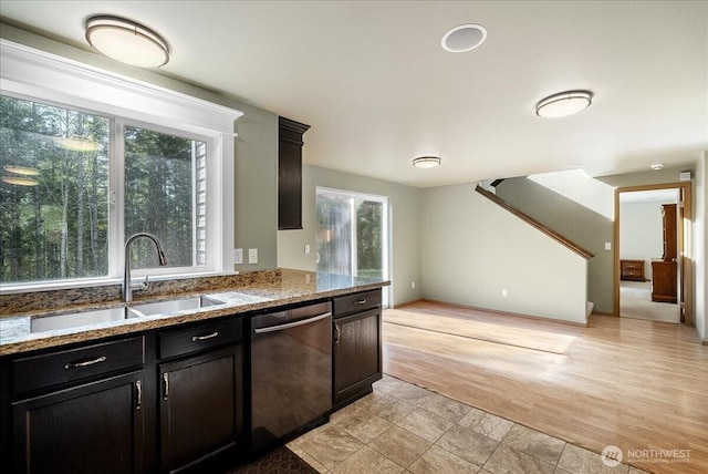 kitchen with light wood-style flooring, light stone countertops, dark cabinetry, stainless steel dishwasher, and a sink