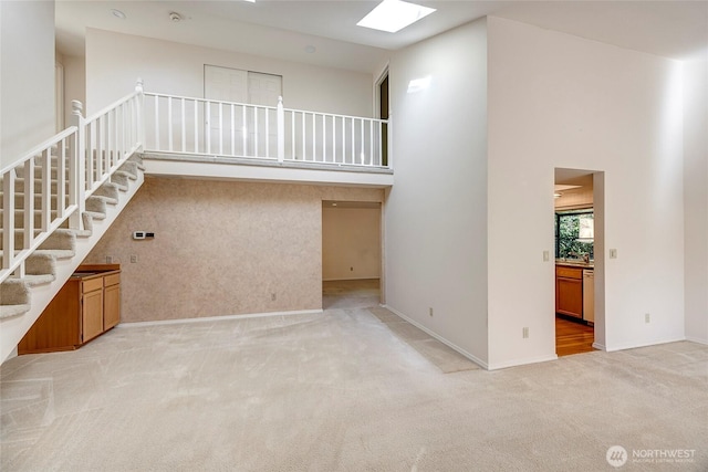 unfurnished living room featuring light carpet, a skylight, a towering ceiling, baseboards, and stairway