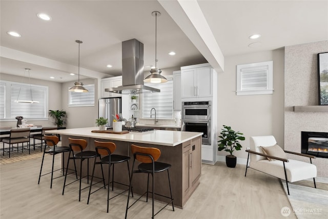 kitchen with a fireplace, a center island with sink, appliances with stainless steel finishes, island range hood, and a kitchen breakfast bar